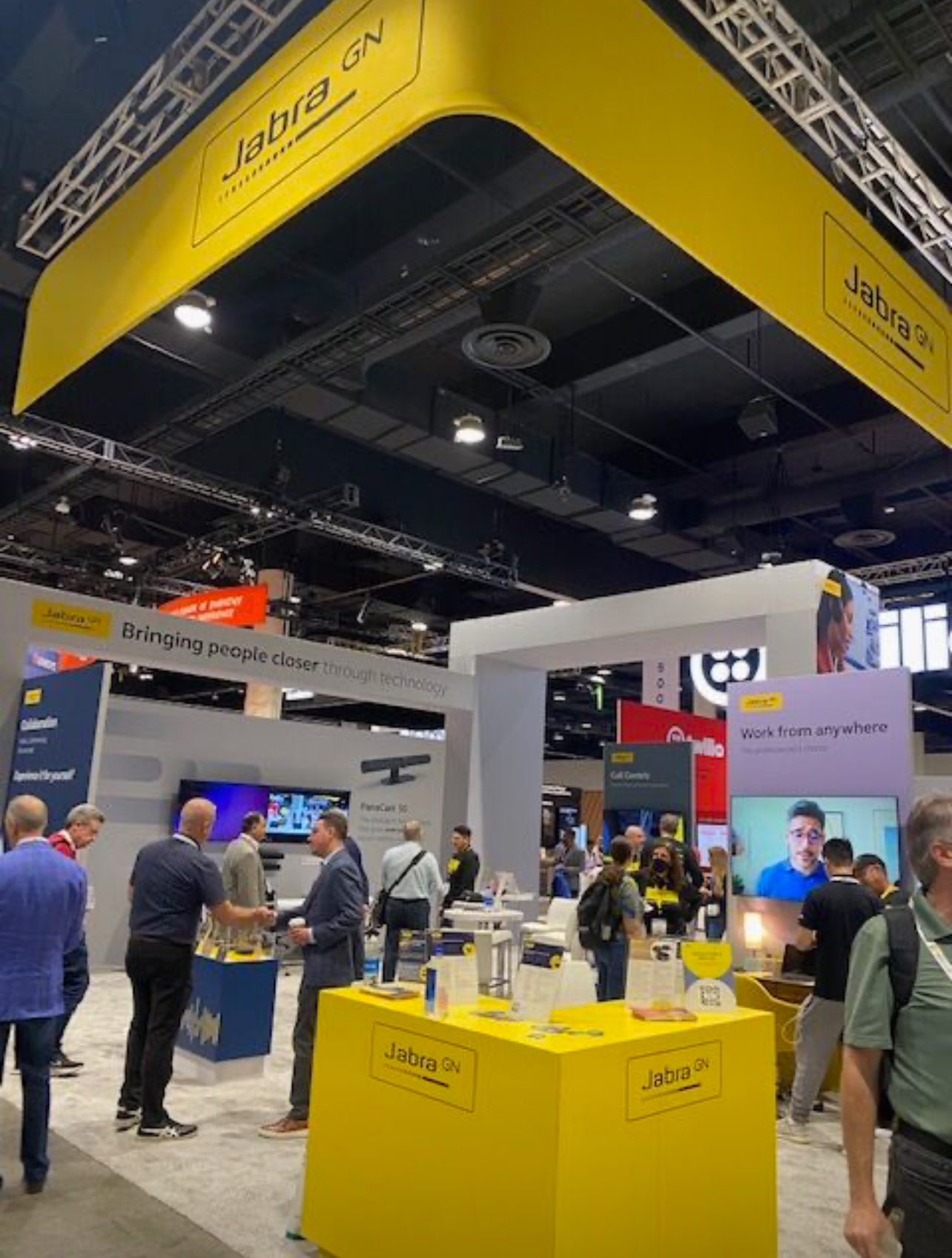 The finished Jabra booth on a convention floor: a yellow overhead banner above white display walls, with attendees talking at the counters.