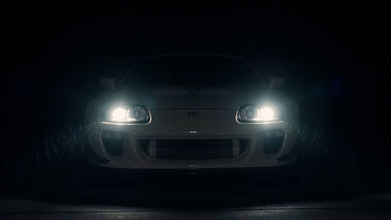 A white Toyota Supra photographed head-on in darkness, headlights lit, the body falling away into black.